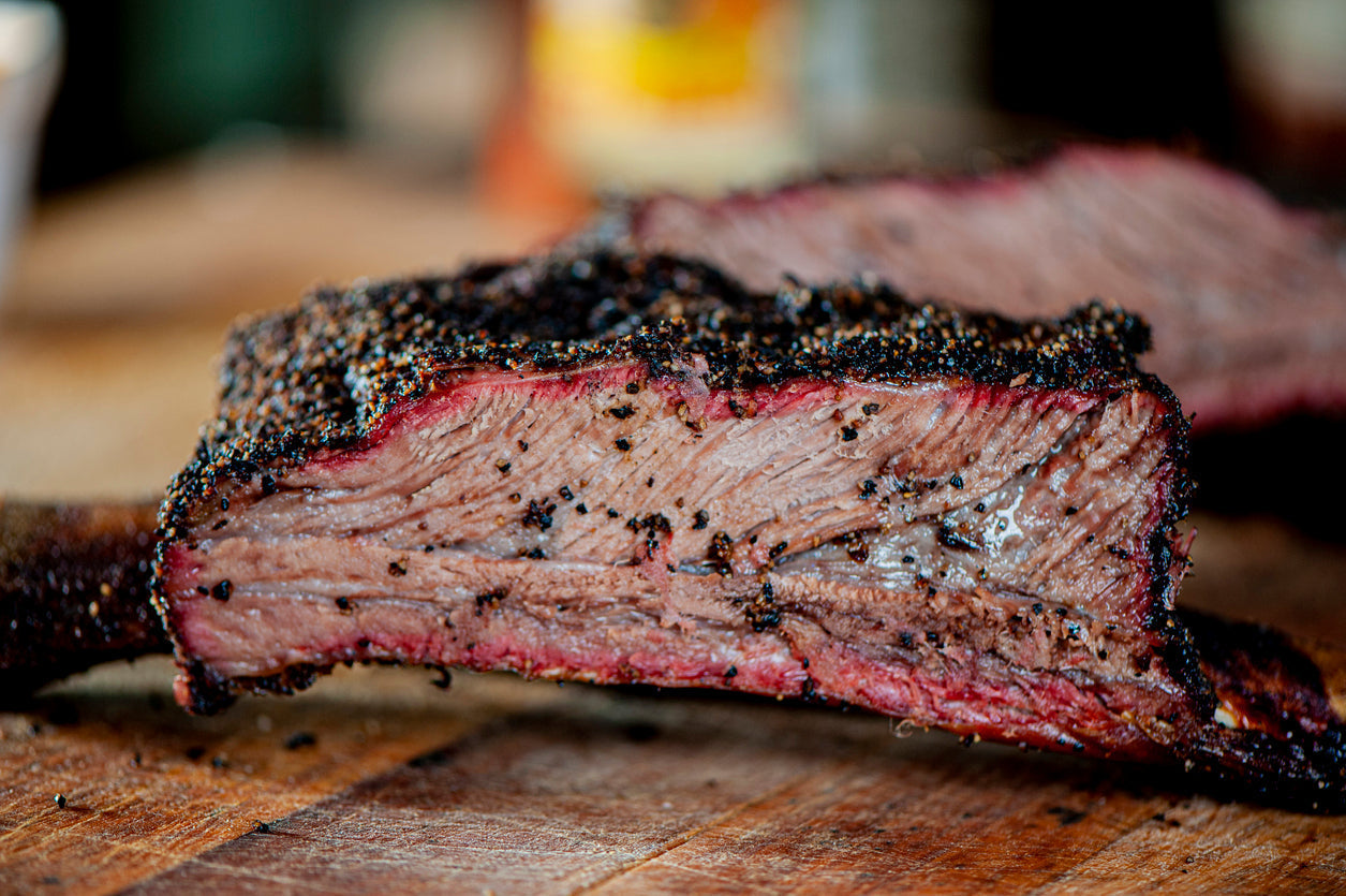 Texas-style smoked brisket sliced on butcher paper with dark bark, visible smoke ring, and rendered juices beside a rustic BBQ setup.