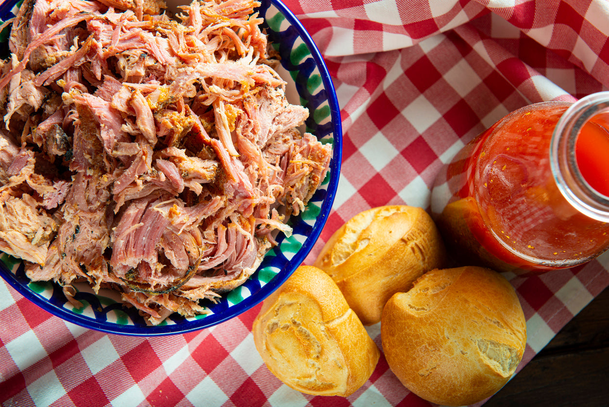 Smoked pulled pork Boston butt shredded on a cutting board with dark bark, BBQ sauce, and smoke visible beside a backyard smoker.